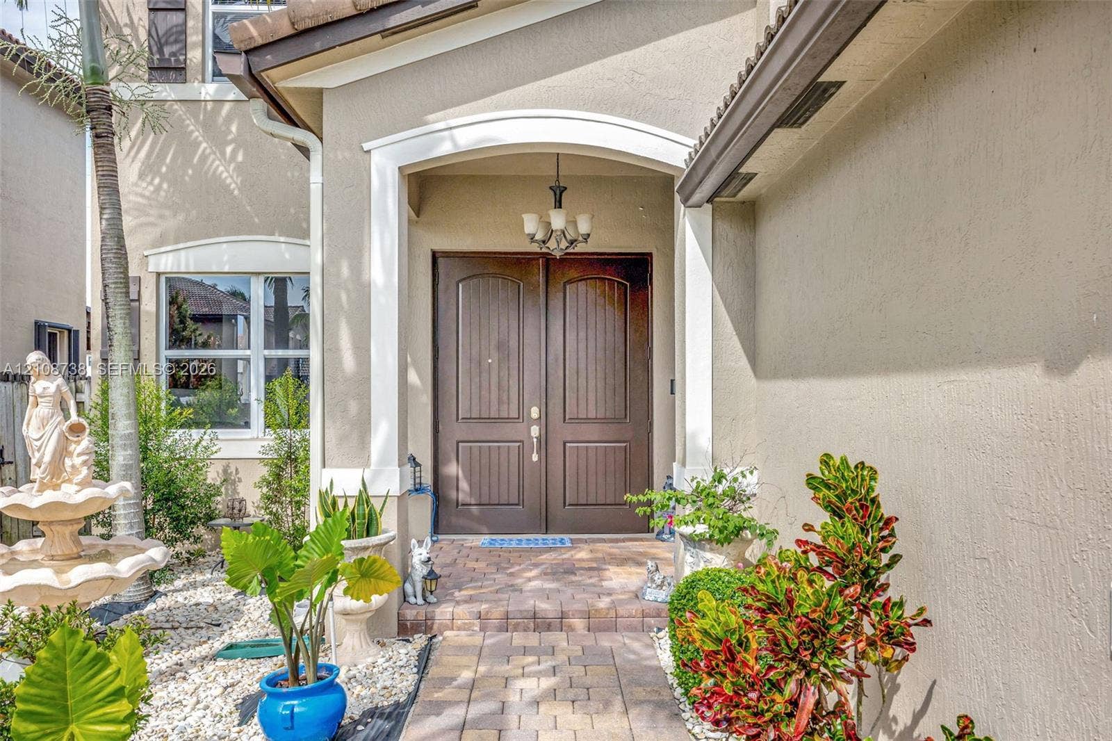 Covered entryway featuring arched architectural detailing, double-door entrance, stone-paved walkway, and a multi-tiered tiered fountain