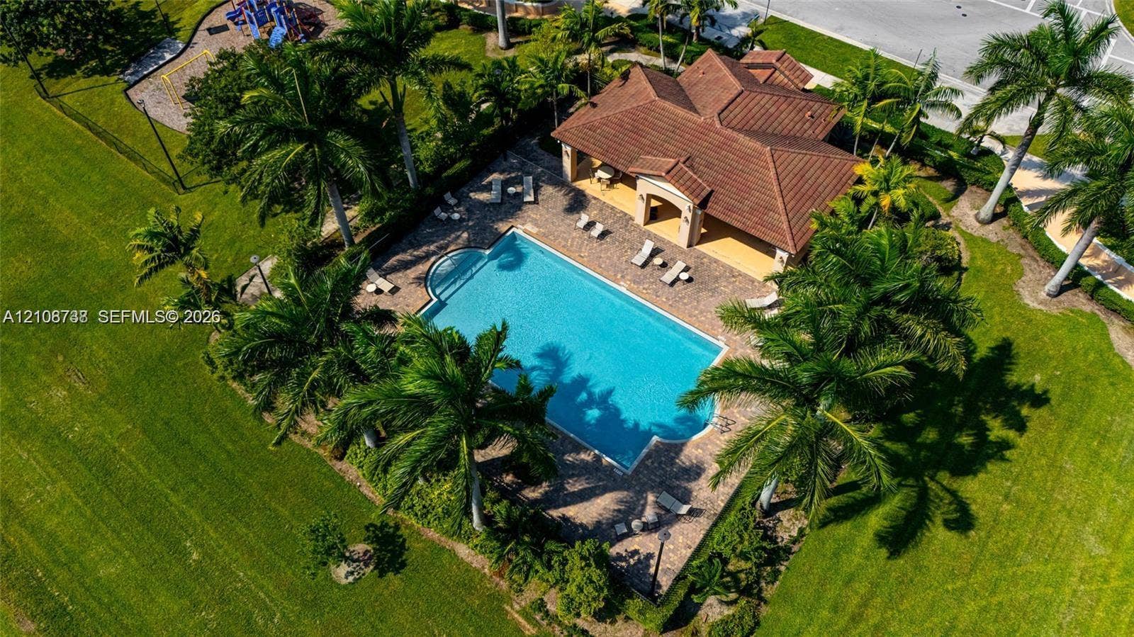 Swimming pool with surrounding paver deck, covered patio area, tile roof structure, and perimeter landscaping featuring mature palm trees