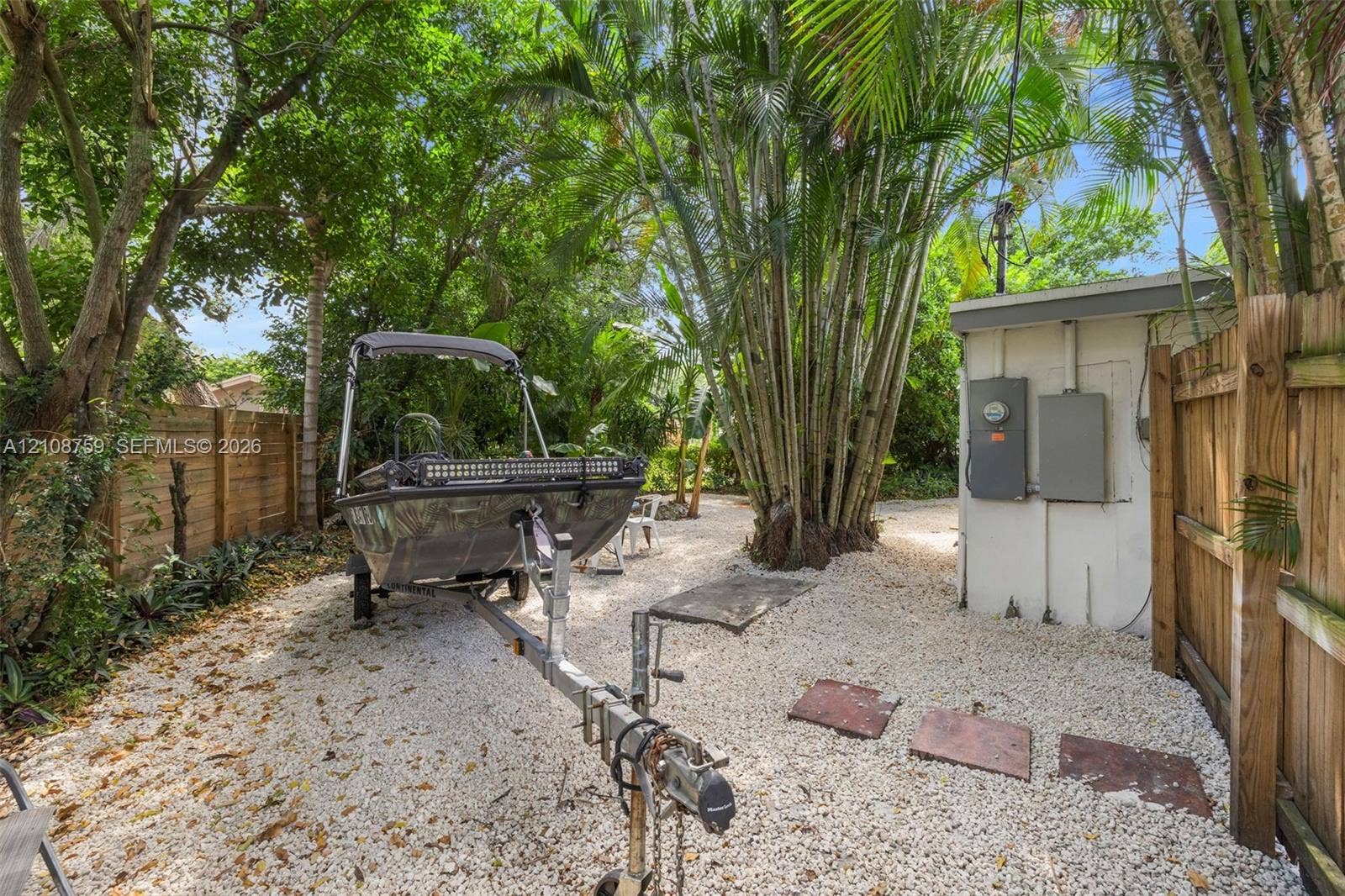 Side yard featuring stone ground cover, vertical wood fencing, mature palm trees, and exterior electrical utility panels
