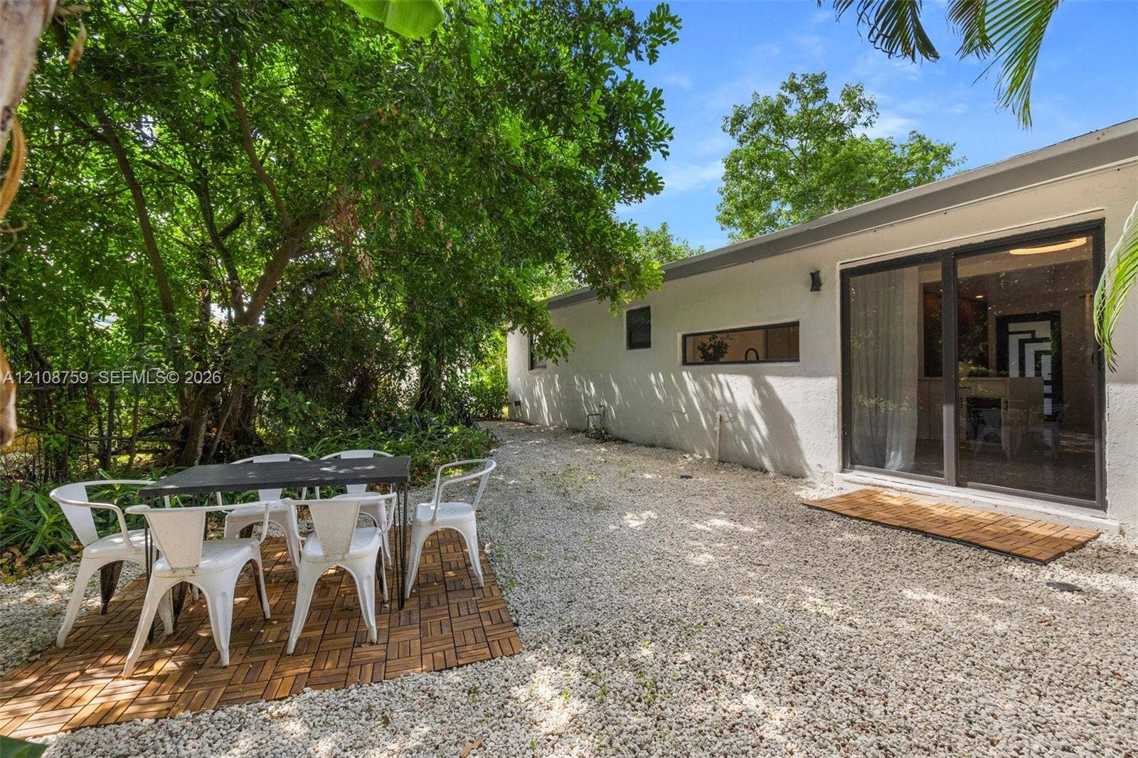 Exterior space featuring light-colored stucco siding, gravel ground covering, sliding glass doors, and mature trees providing shade