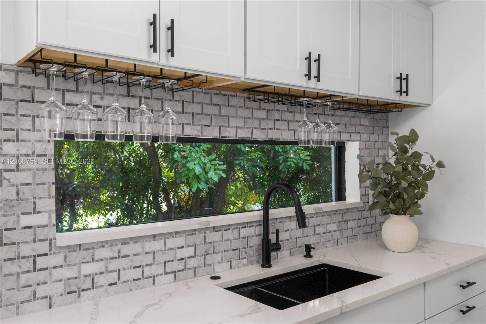 White Shaker-style cabinetry with built-in stemware racks, stone-tiled backsplash, quartz countertops, undermount double-basin sink with a matte black pull-down faucet, and a horizontal window
