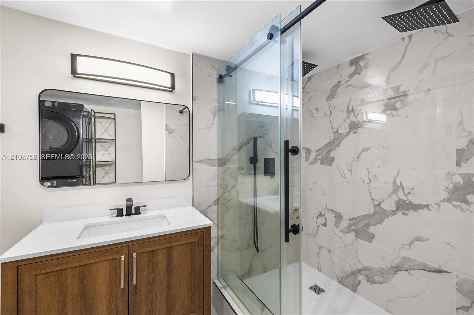 Bathroom featuring a wood-finish vanity with a white countertop, large wall-mounted mirror, sliding glass shower door, and rainfall showerhead with stone-patterned wall tile