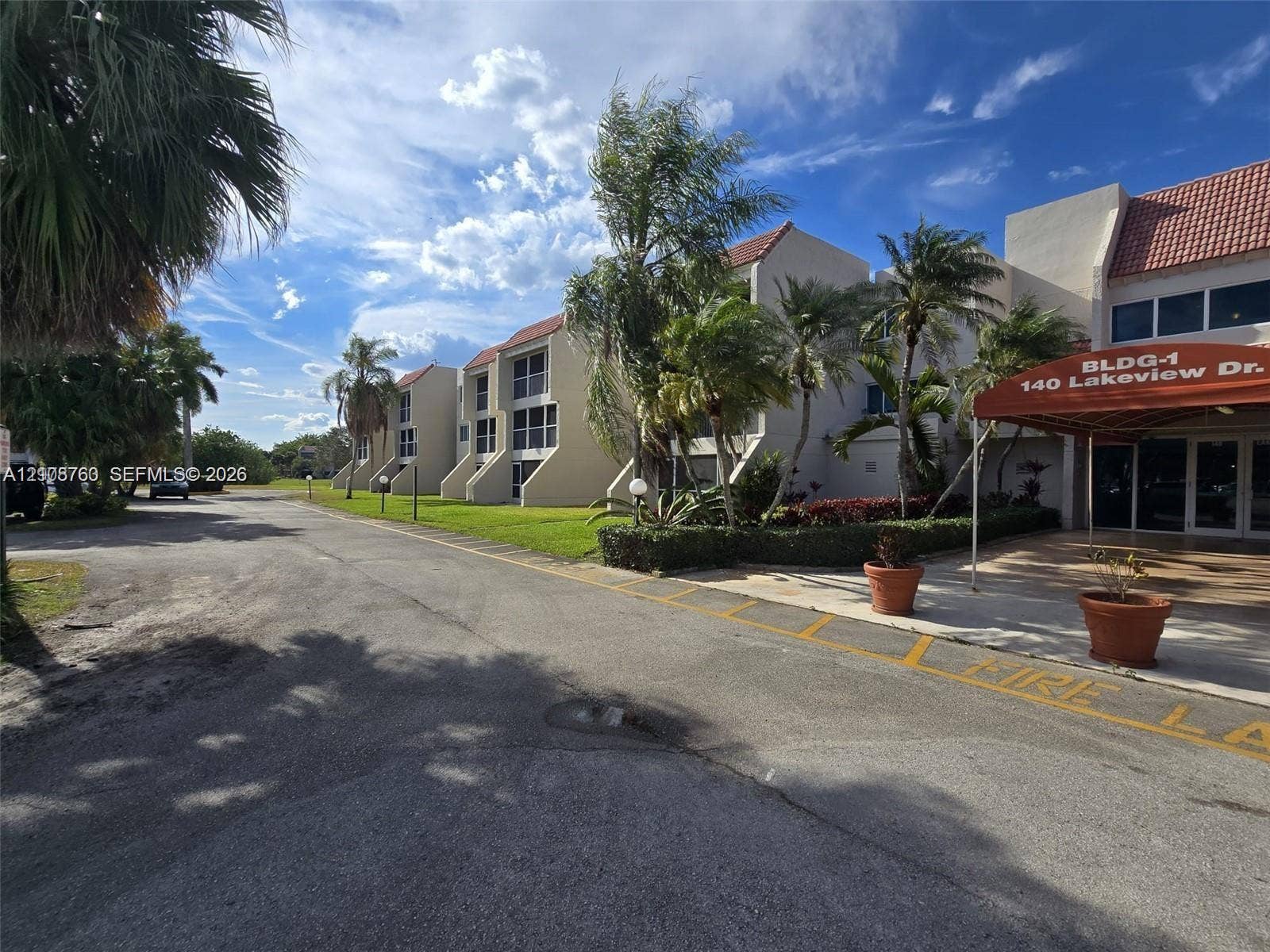 140 Lakeview Drive, #111, Weston, Weston, FL 33326 Multi-story exterior featuring a light-toned stucco faade, terra-cotta-hued roof tiles, and ground-level covered entryway with a red fabric awning, complemented by landscaped perimeter gardens and mature palms
