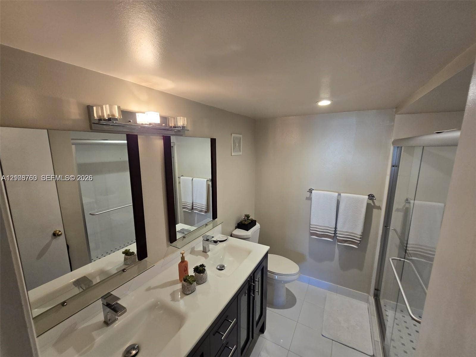 140 Lakeview Drive, #111, Weston, Weston, FL 33326 Dual-sink vanity with a solid-surface countertop, dark cabinetry, full-width mirror, and a walk-in glass-enclosed shower featuring tiled flooring
