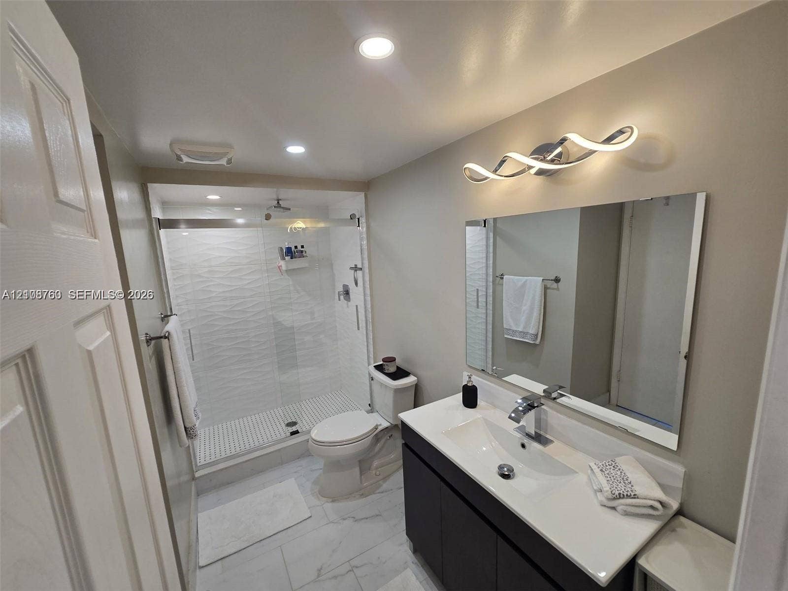 140 Lakeview Drive, #111, Weston, Weston, FL 33326 Bathroom featuring a dark wood-finish vanity with an integrated countertop sink, walk-in shower with textured wall tile, frameless glass enclosure, and contemporary wave-style wall lighting