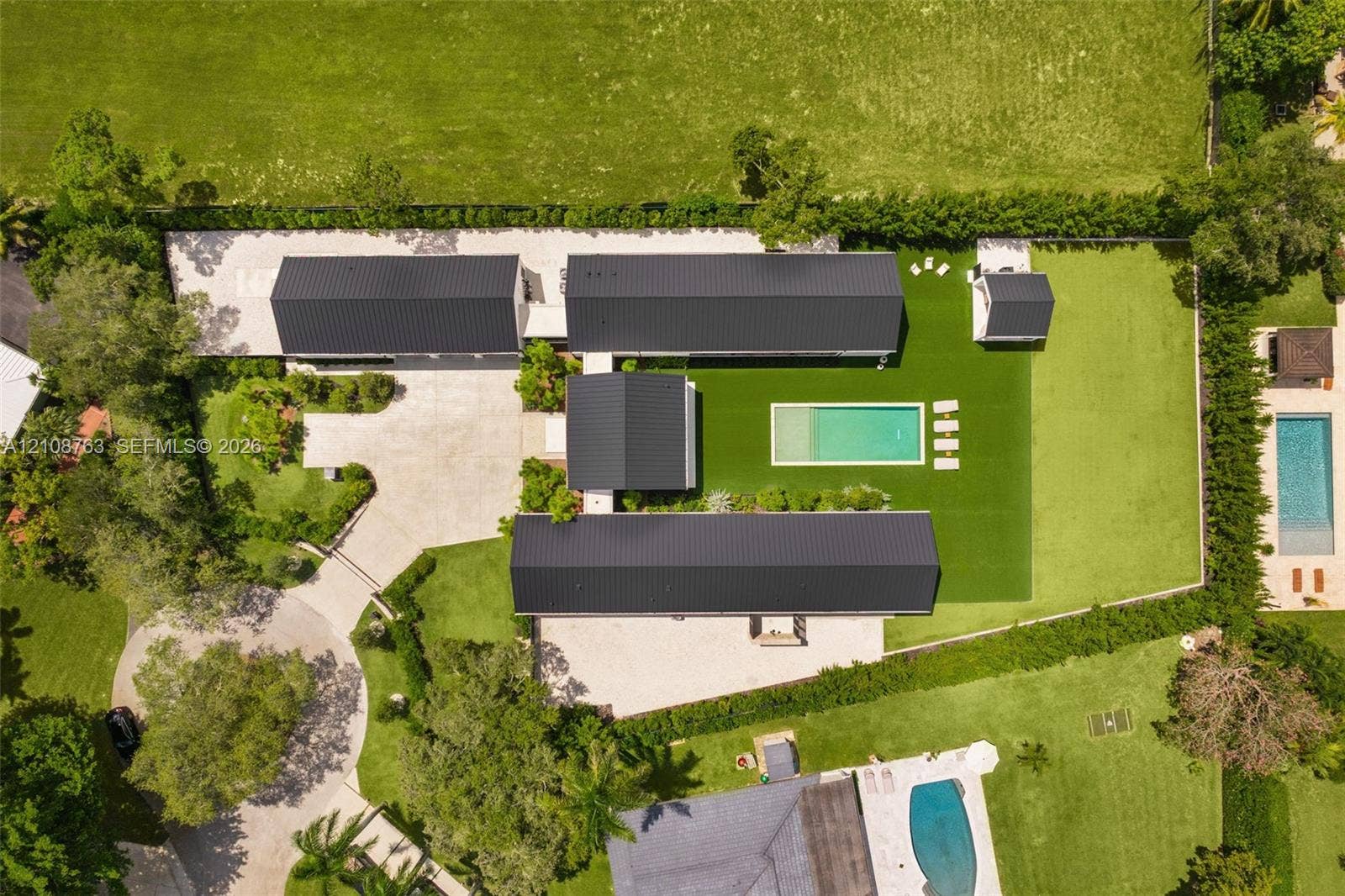 Aerial perspective of a multi-wing residence featuring a dark metal roof, rectangular swimming pool, manicured lawn, and extensive paved driveway