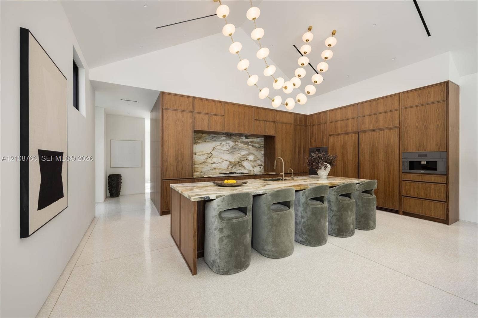 Modern kitchen featuring full-height wood cabinetry, a stone-top island with integrated sink, polished light-colored flooring, vaulted ceiling, and tiered orb pendant lighting