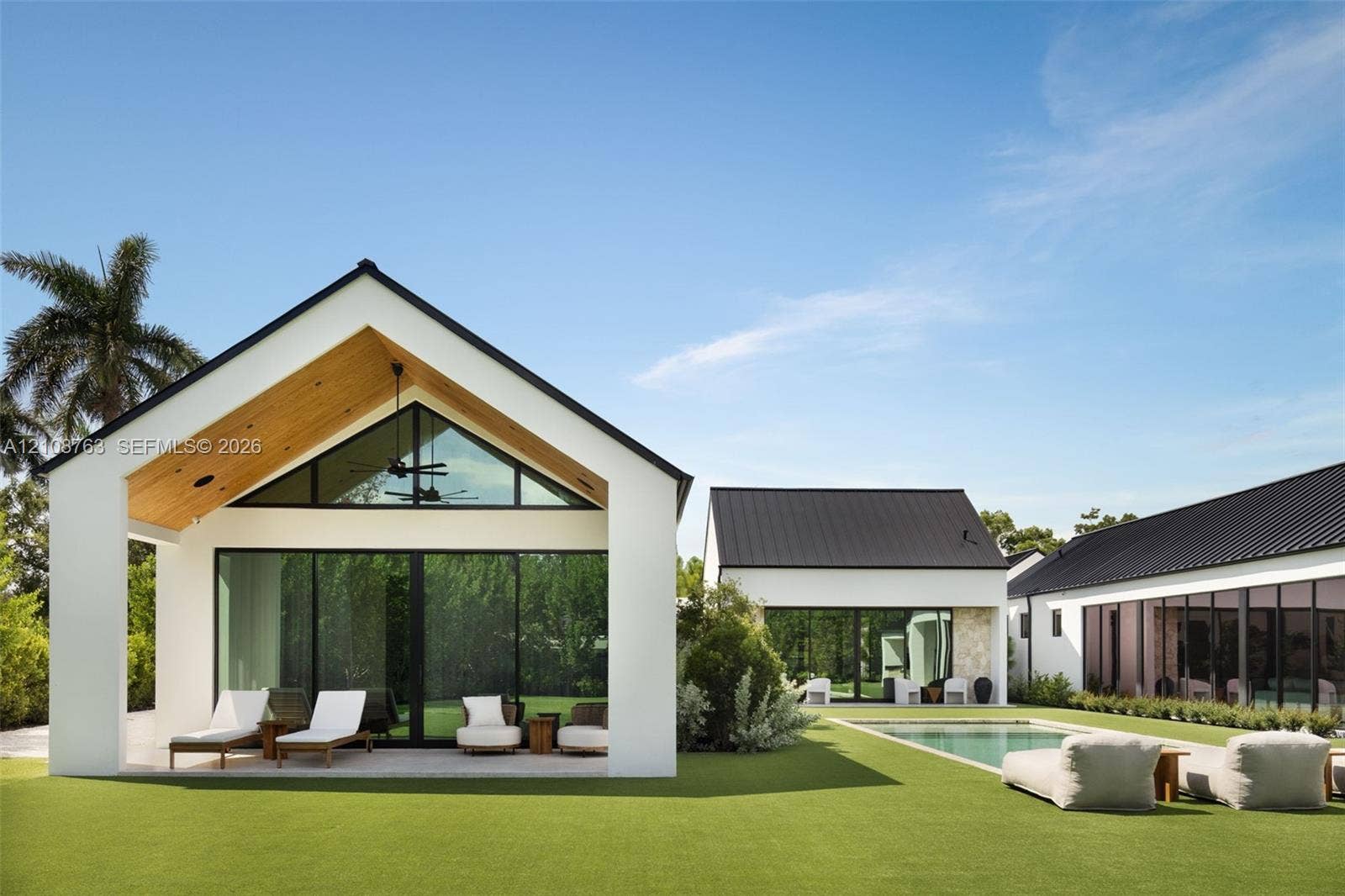 Contemporary exterior featuring a gabled roofline, light-toned wood-finish ceiling under the covered patio, expansive floor-to-ceiling glass panel walls, dark-toned metal roofing, and a rectangular swimming pool with stone hardscaping