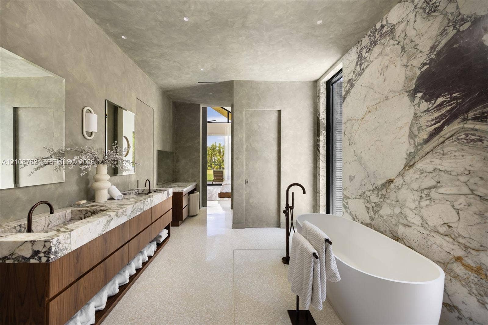 Custom vanity with integrated stone sinks and wood-finish cabinetry, freestanding soaking tub, expansive stone-slab feature wall, and recessed ceiling lighting