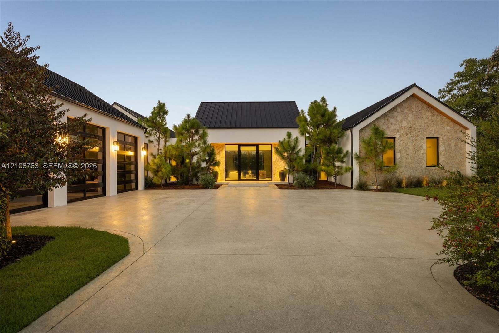 Modern estate featuring a standing-seam metal roof, smooth stucco finish, stone-clad exterior wing, and expansive concrete motor court with multi-car garage glass-panel doors
