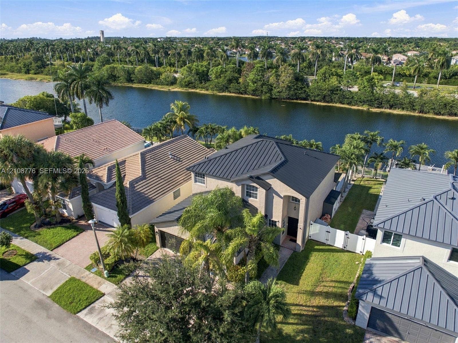 18041 SW 33rd Street, Miramar, FL 33029 Stucco exterior residence featuring a metal roof, paved driveway, white vinyl perimeter fencing, and water frontage