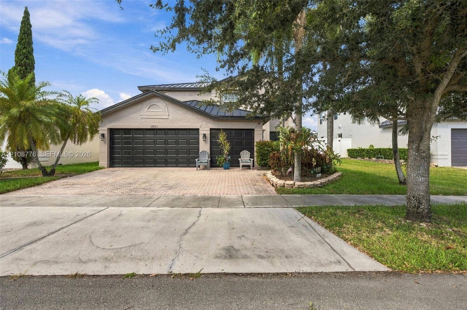 18041 SW 33rd Street, Miramar, FL 33029 Multi-level exterior featuring a metal roof, paver driveway, three-car garage with dark-finish doors, and landscaped garden beds