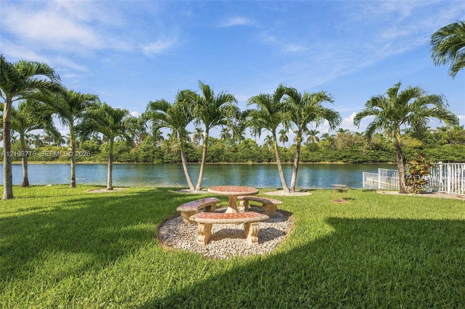 18041 SW 33rd Street, Miramar, FL 33029 Waterfront backyard featuring a manicured lawn, palm trees, and a decorative stone-finish seating area set on a gravel bed