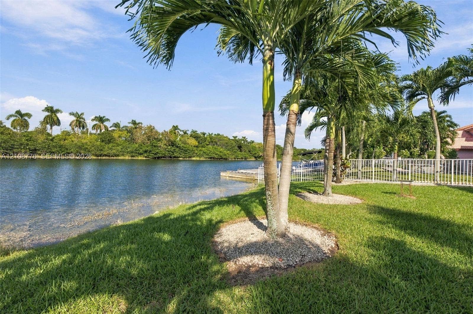18041 SW 33rd Street, Miramar, FL 33029 Waterfront grounds featuring a grass lawn, mature palm trees, decorative stone tree rings, and a perimeter fence