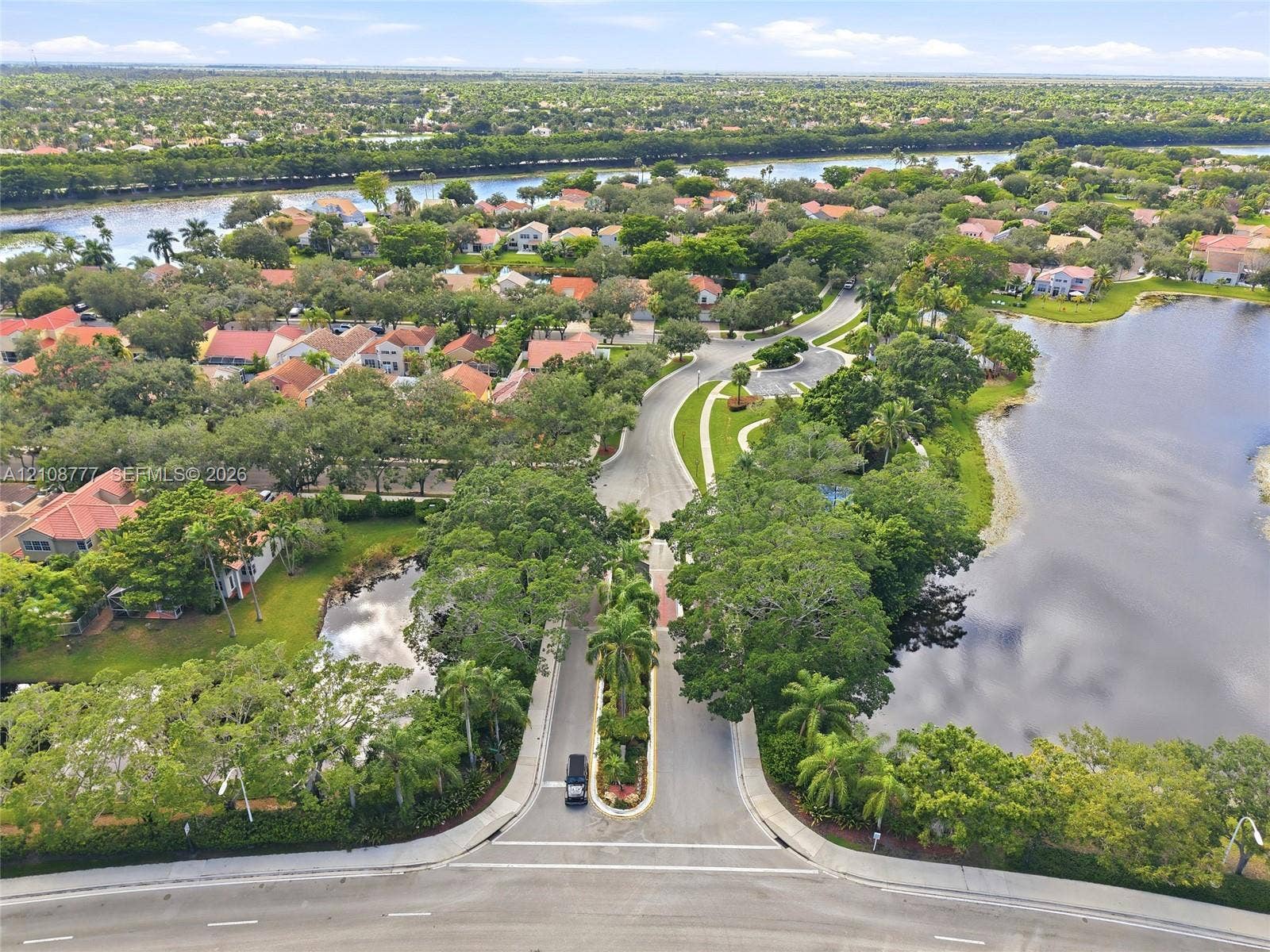 Aerial perspective of a community entrance featuring paved roadways, mature tree canopy, waterfront borders, and landscaped median
