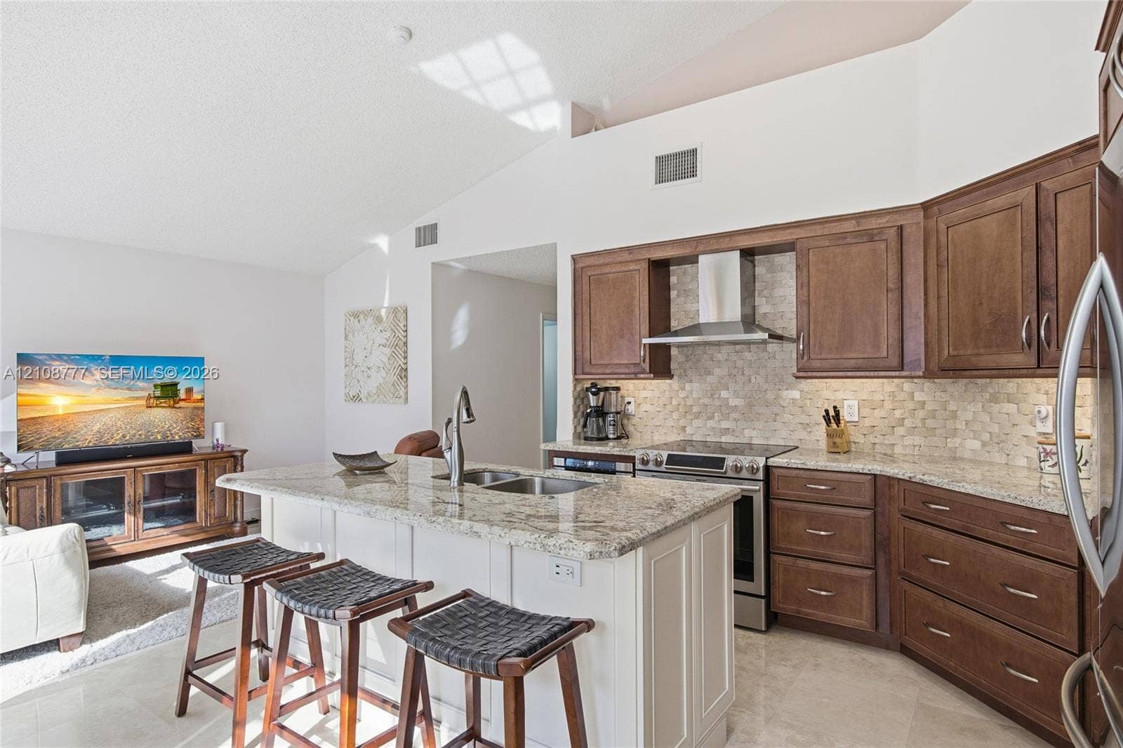 Kitchen featuring custom wood cabinetry, stone countertops, stone-tile backsplash, stainless steel range hood, and a central island with a built-in sink