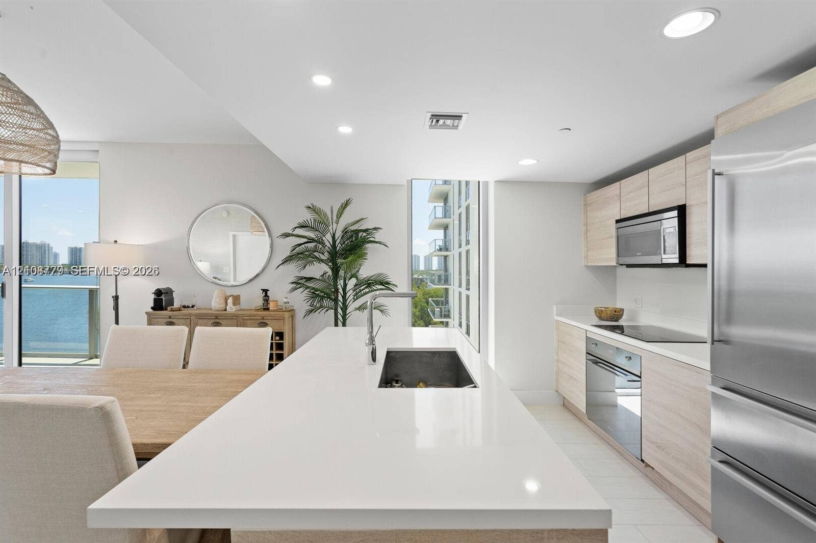 Modern kitchen featuring a central island with a solid-surface countertop and undermount sink, flat-panel light wood-finish cabinetry, stainless steel appliances, recessed ceiling lighting, and large-format tile flooring