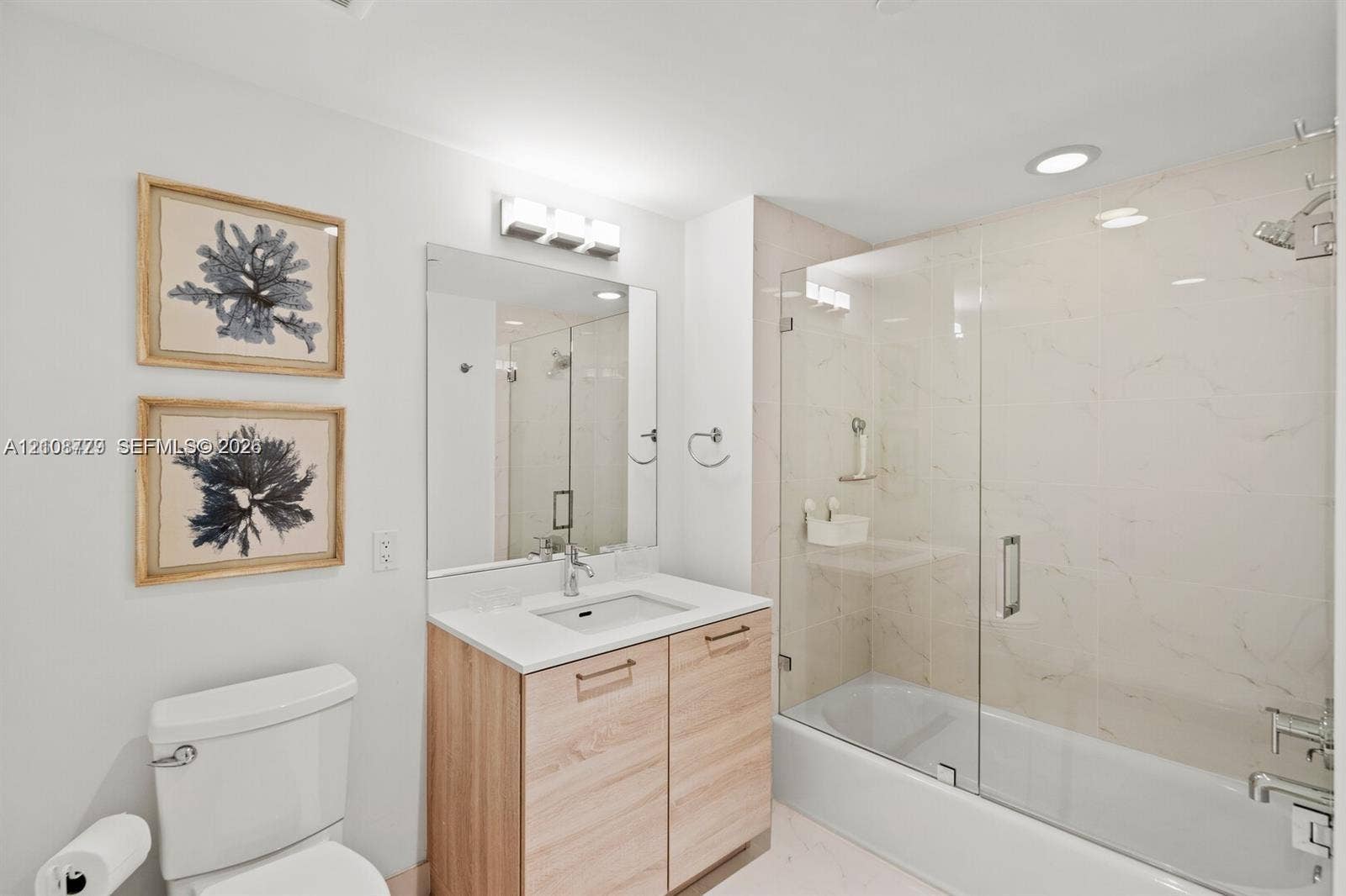 Bathroom featuring a wood-finish vanity with a rectangular sink, stone-surface countertop, full-height wall mirror, and a tub-shower combination enclosed by a frameless glass door with light-toned wall tile