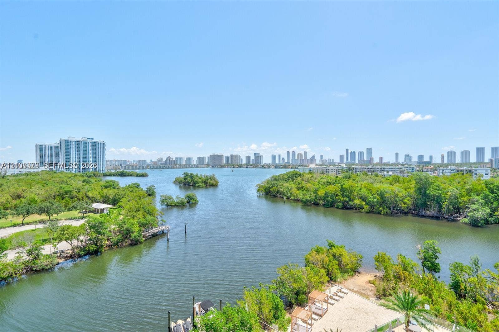 Waterfront locale featuring mangrove-lined shorelines, a sandy recreation area with shaded structures, and a distant cityscape skyline