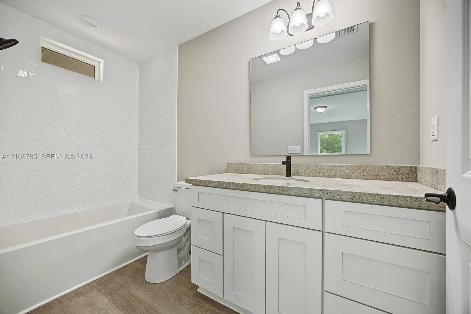 Bathroom featuring a vanity with white shaker-style cabinetry, neutral stone-finish countertop, undermount sink with dark metal hardware, and wood-finish flooring, complemented by a tiled shower surround