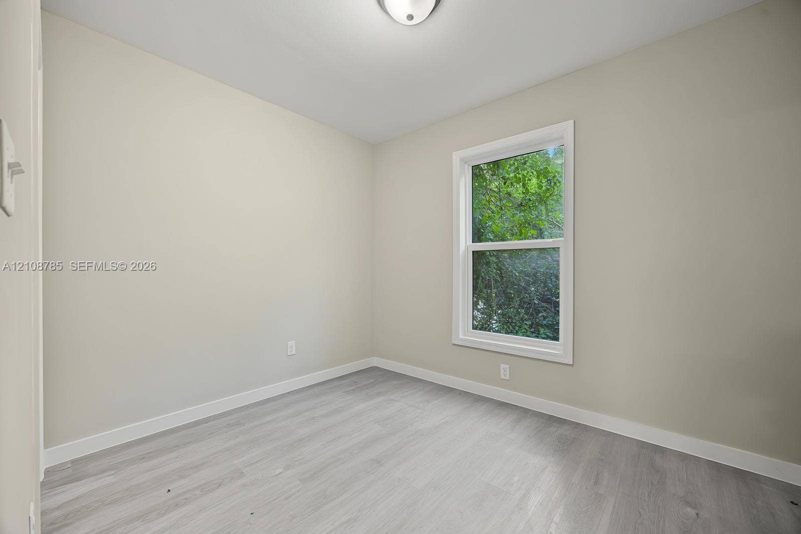 Neutral-toned walls, wood-finish flooring, and a double-hung window with a verdant exterior outlook, illuminated by a ceiling-mounted light fixture