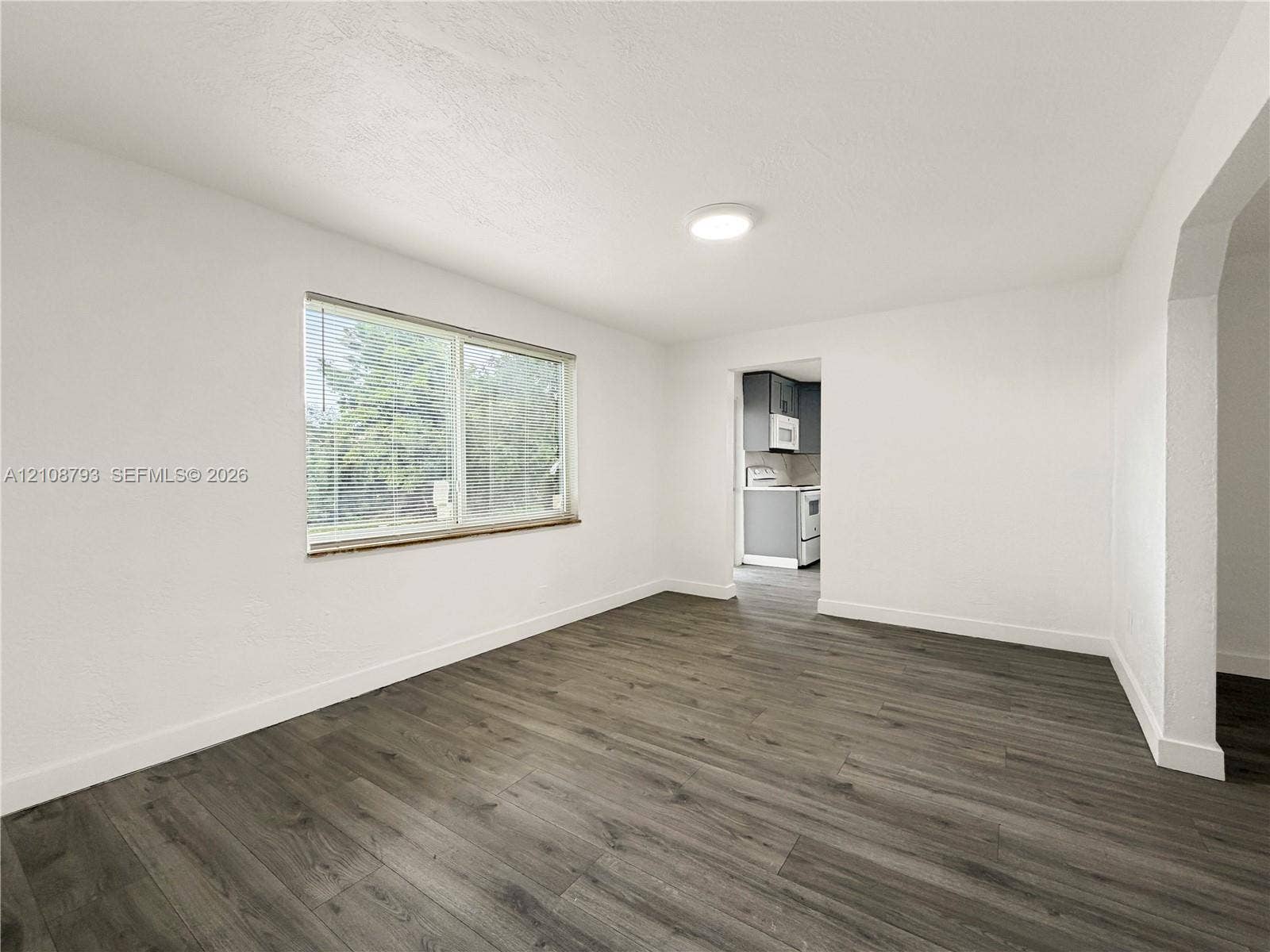 Neutral-toned interior featuring wood-finish flooring, white walls, flush-mount ceiling lighting, a large window with horizontal blinds, and a wide arched opening