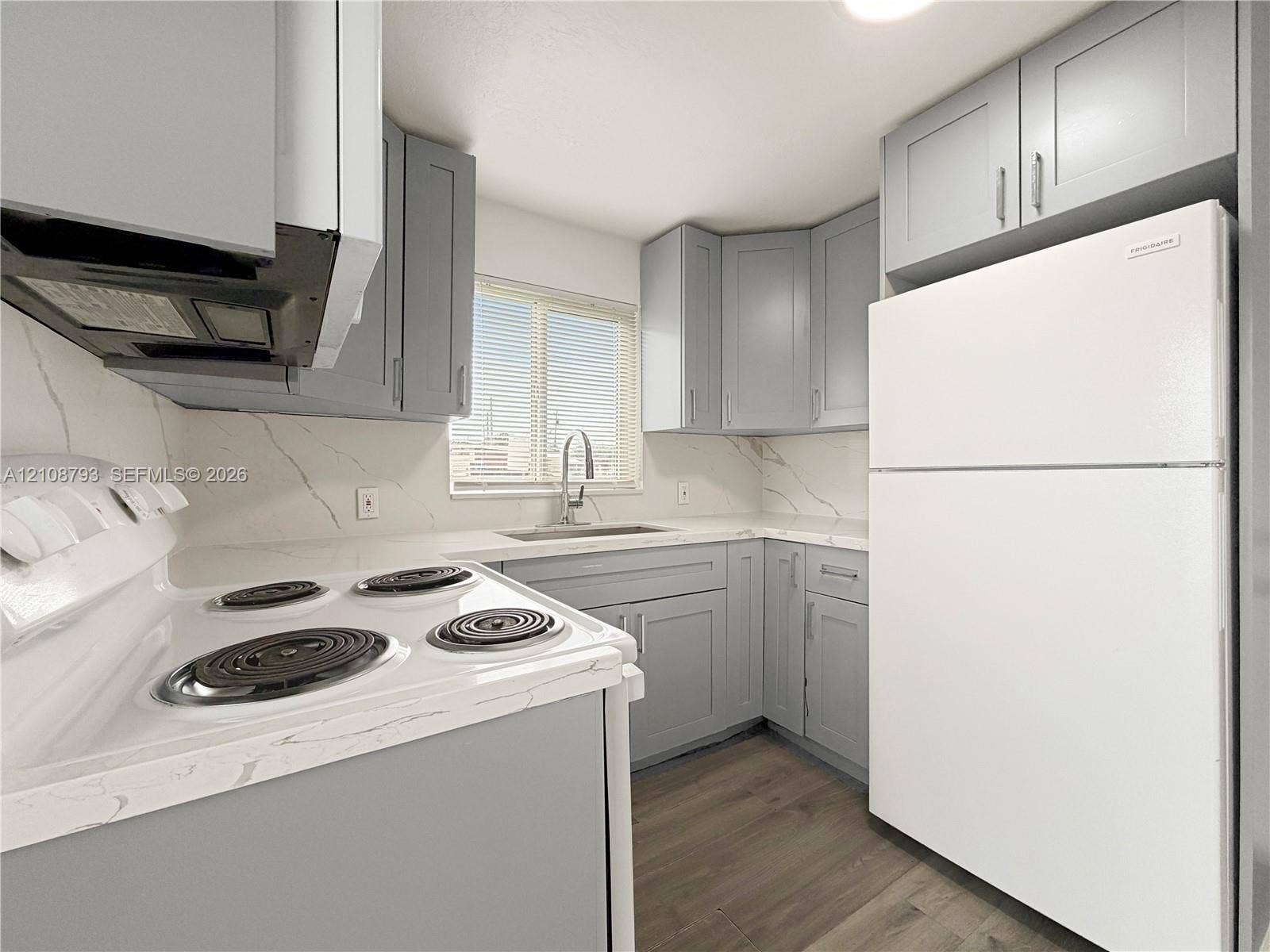 Kitchen featuring shaker-style cabinetry, white countertops with veining, full-height backsplash, wood-finish flooring, and a stainless steel kitchen sink