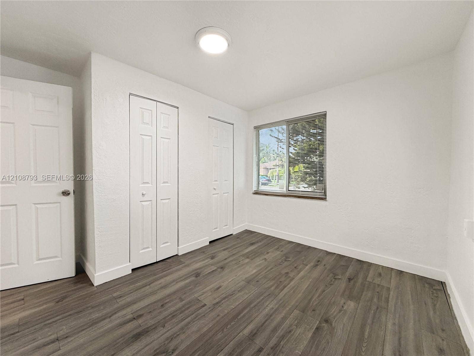 Neutral-toned room featuring wood-finish flooring, white walls, two sets of paneled closet doors, and a sliding window with metallic framing