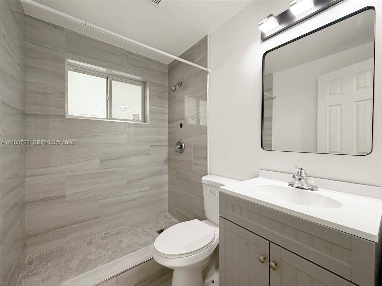 Walk-in shower featuring large-format tile walls, integrated hexagonal floor tiling, grey cabinetry with a white countertop, and a wall-mounted rectangular mirror