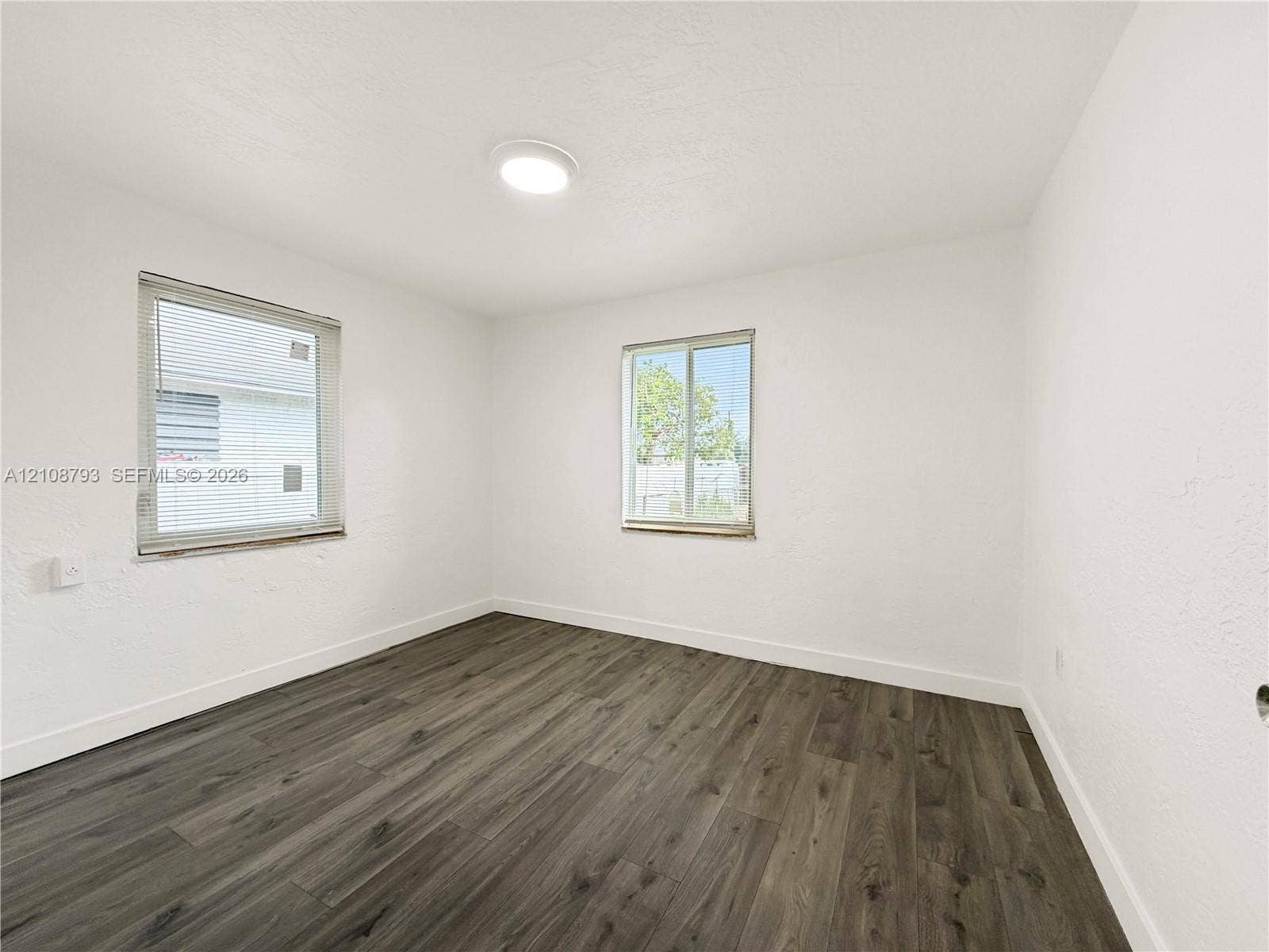 Room featuring wood-finish flooring, two windows with blinds, and a flush-mount ceiling light fixture