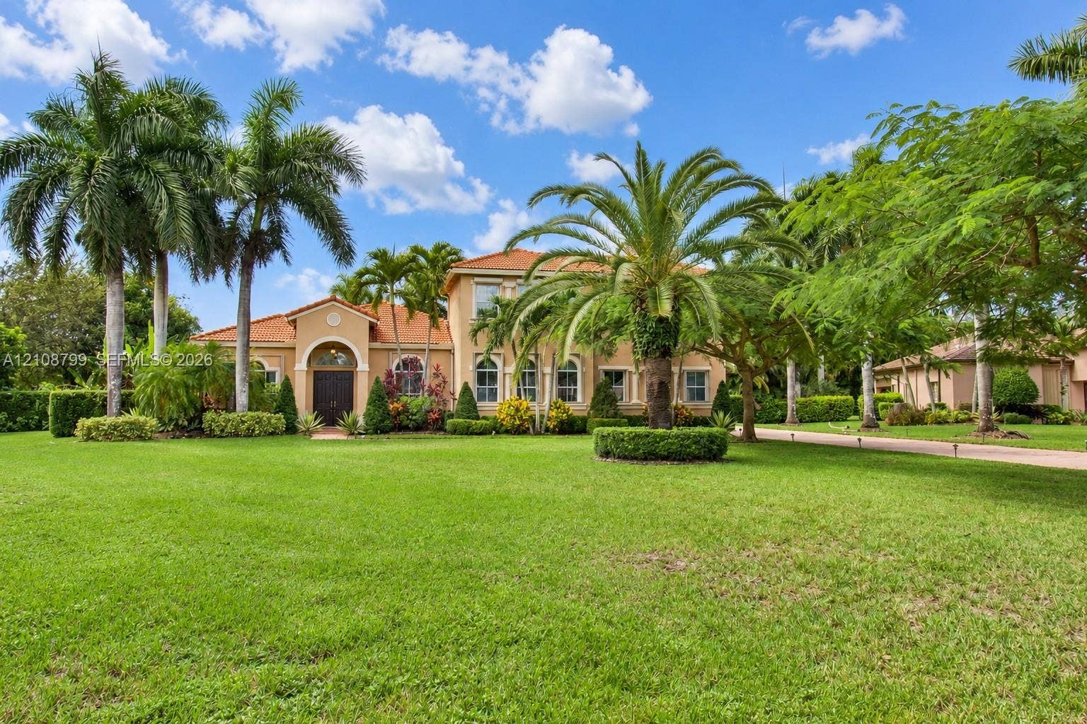 Stucco exterior with an arched entryway, terracotta tile roof, and expansive manicured lawn featuring mature palm trees
