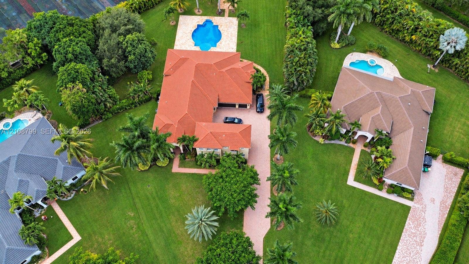 Aerial perspective featuring a multi-gabled residence with a terracotta tile roof, an expansive landscaped lawn, a private swimming pool with concrete decking, and a paved driveway