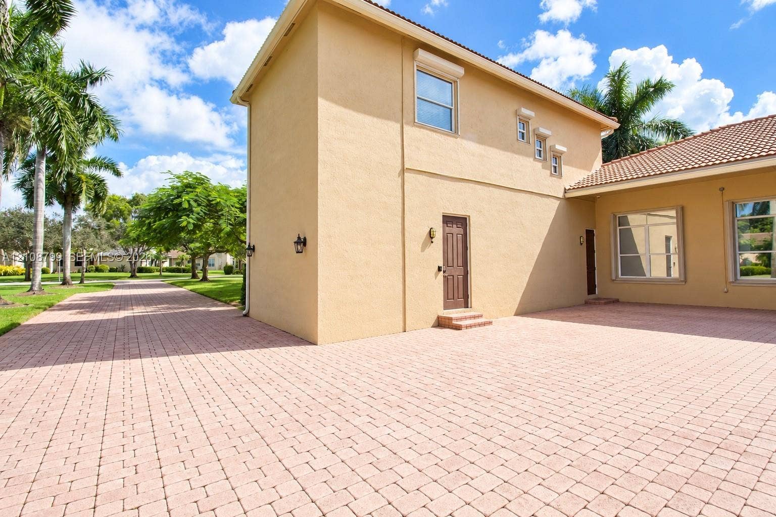 Stucco exterior featuring an expansive paved driveway, a multi-level roofline with clay tile detailing, exterior wall-mounted lighting, and integrated shutters