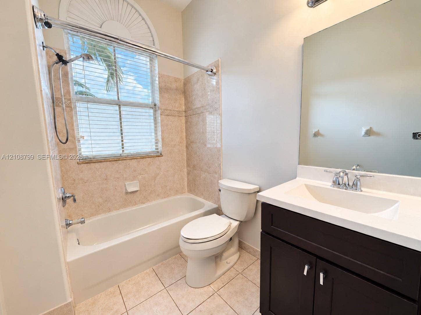 Bathroom featuring a dark wood-finish vanity with a white countertop and integrated sink, a tiled tub-shower combination, ceramic tile flooring, and a window with an arched transom