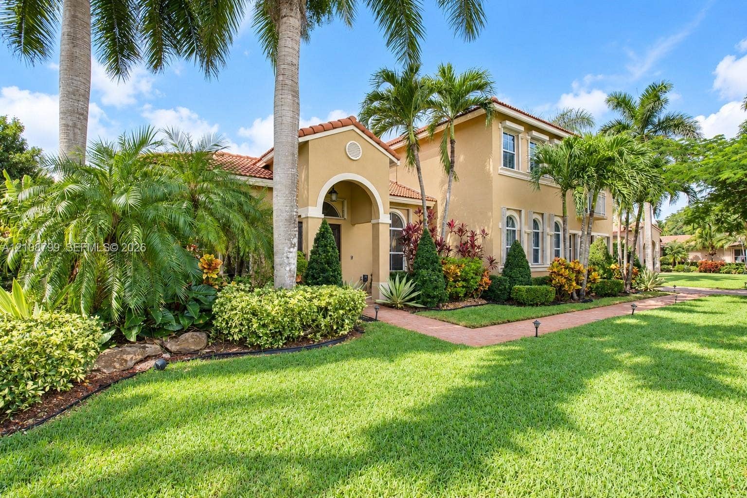 Stucco exterior featuring a tiled roof, arched entryway, and manicured landscaping with mature palm trees and a paved walkway