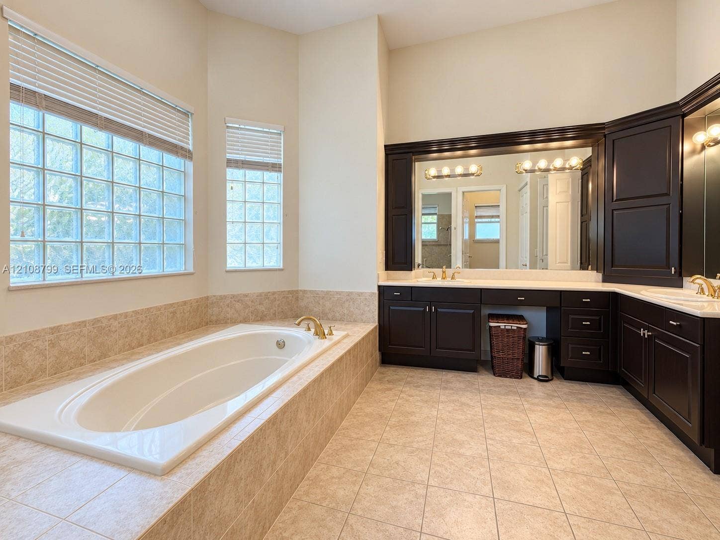 Soaking tub with tile surround, dual-sink vanity with dark cabinetry, light-toned tile flooring, and multi-paned glass block windows
