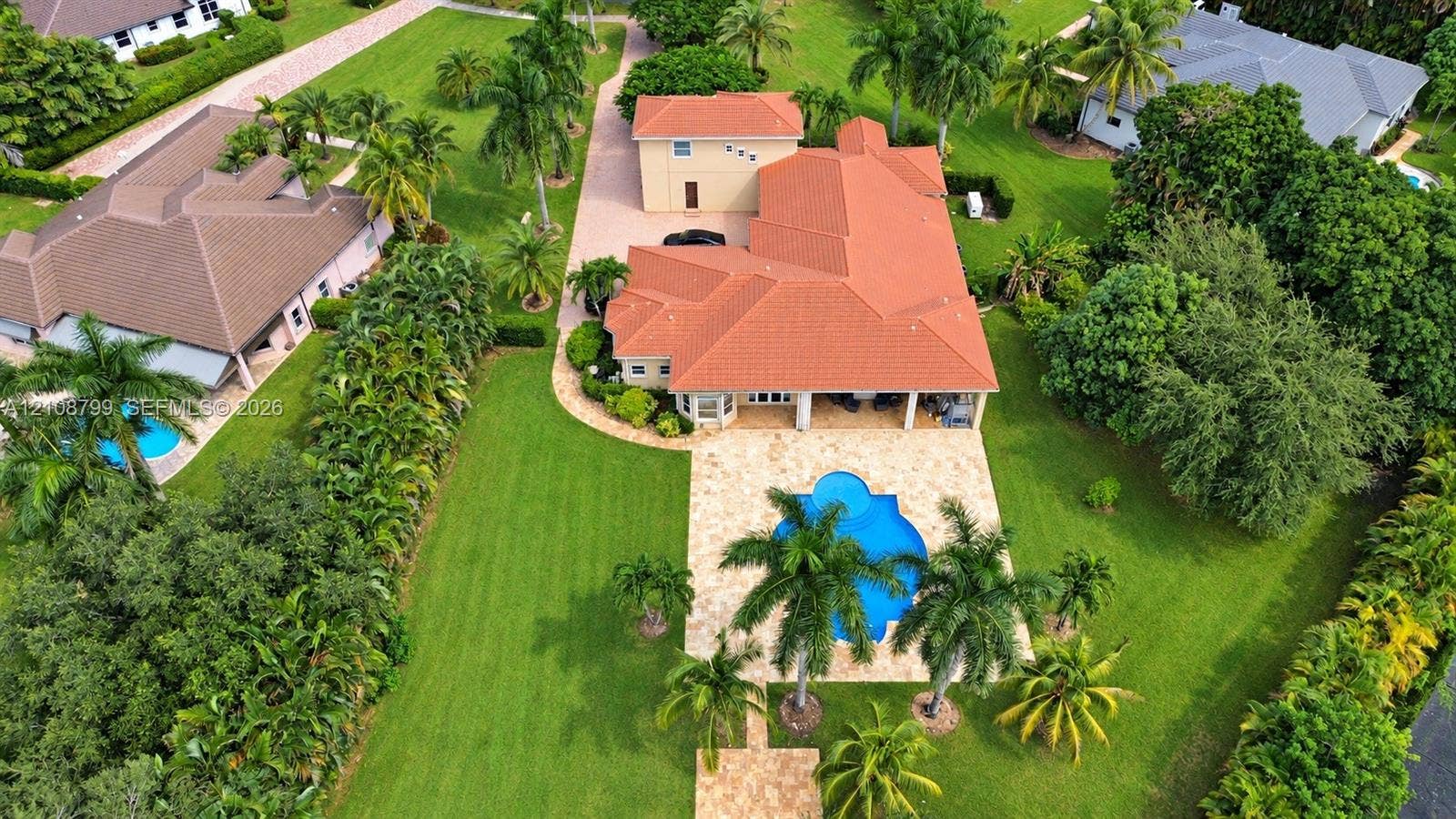 Expansive property featuring a multi-level structure with a terracotta tile roof, a stone-paved patio, a custom-shaped swimming pool, and mature palm trees within a manicured lawn