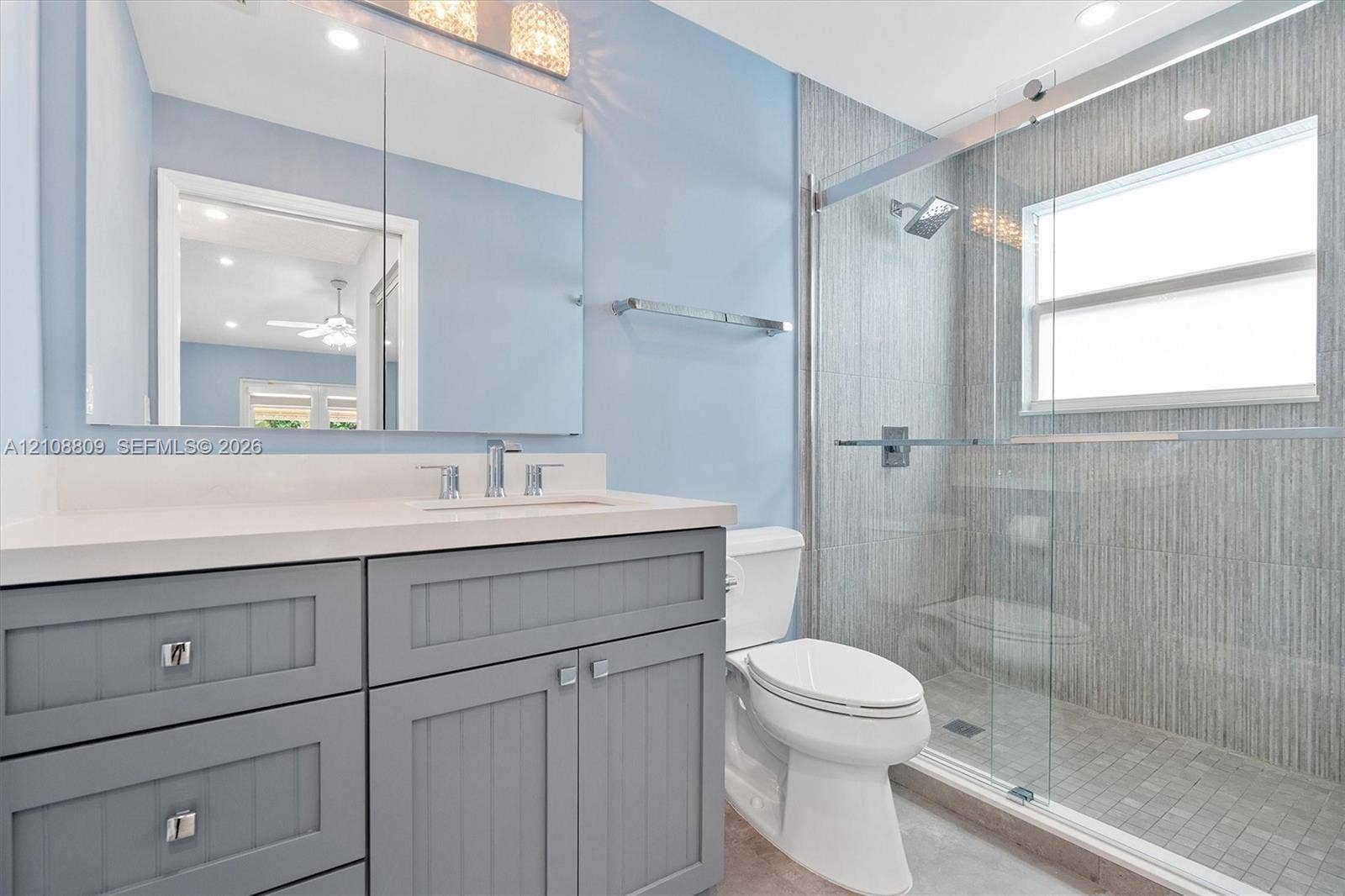 Bathroom featuring a gray vanity with beadboard-style cabinetry, solid-surface countertop, full-width mirror, glass-enclosed shower with textured tile surround, and chrome fixtures