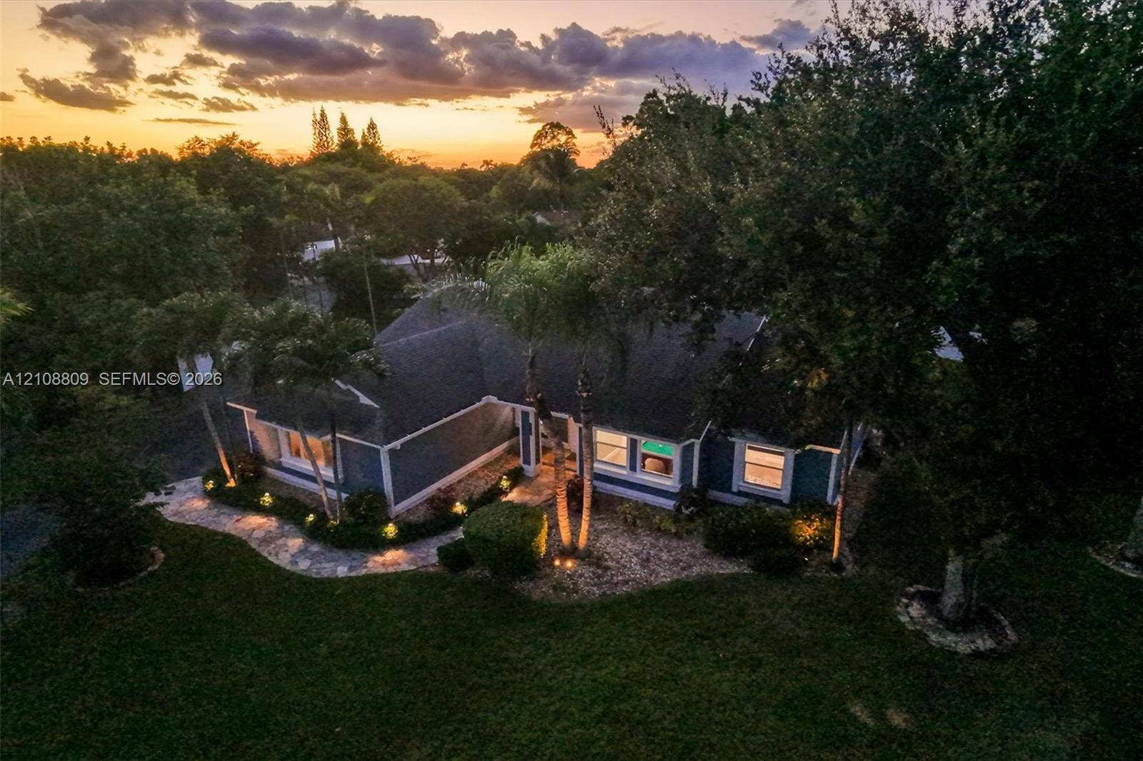 Single-story residence featuring a shingled roof, exterior wall-mounted lighting, manicured landscaping, stone-paved walkway, and mature palms