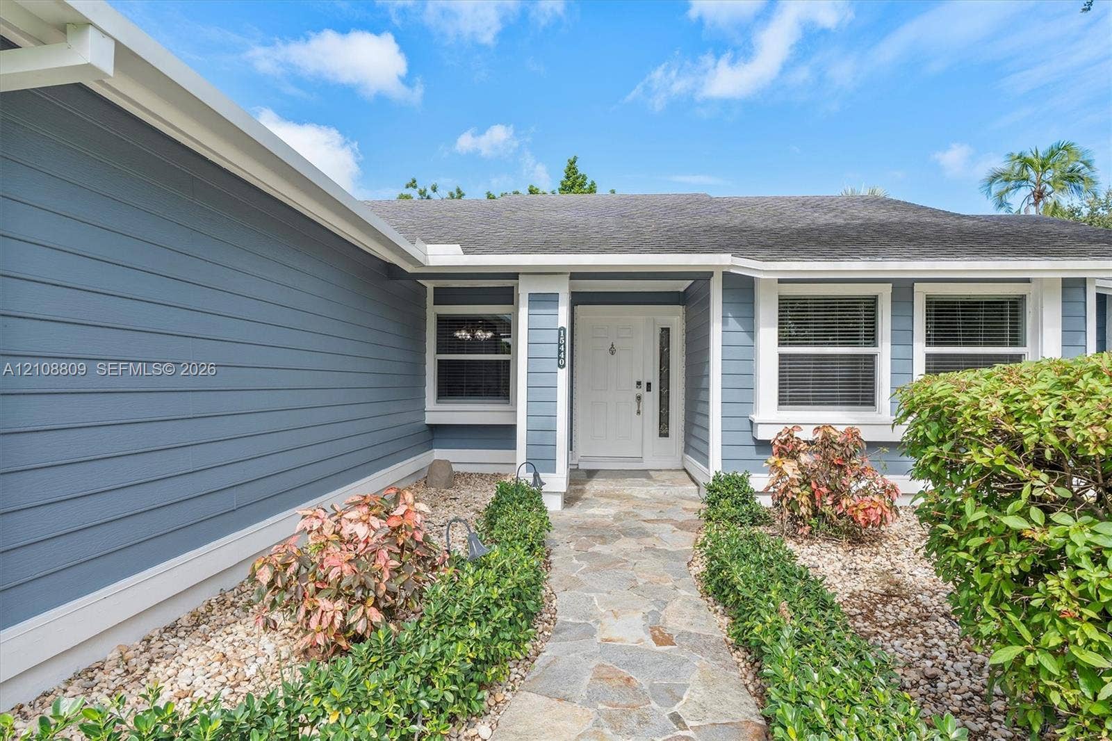 Blue horizontal siding exterior featuring a white-framed entry door with a vertical sidelight, stone paver walkway, and structured landscape hedging