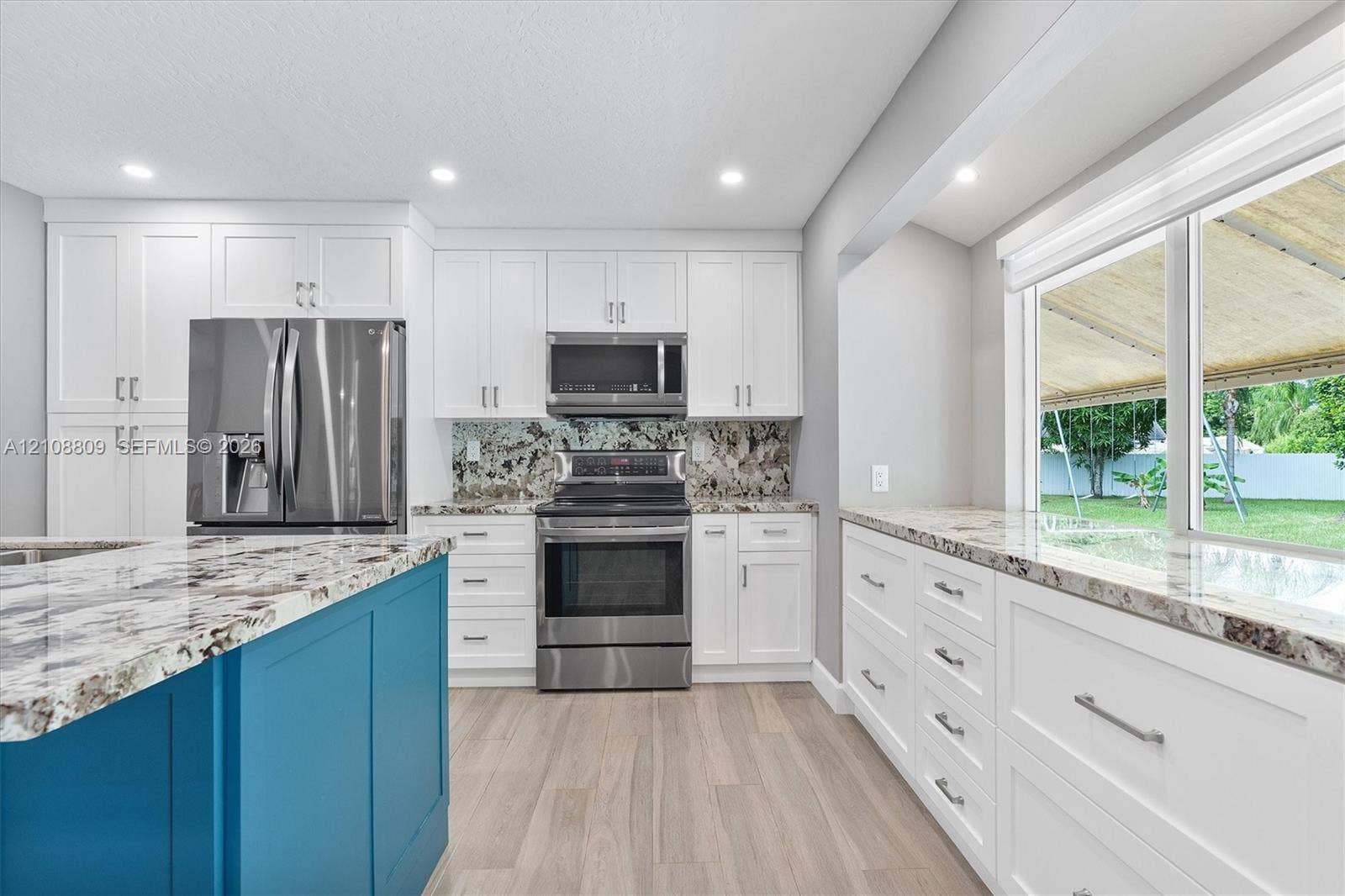 Kitchen featuring white shaker-style cabinetry, a contrasting blue island, patterned stone countertops, stainless steel appliances, and wood-finish flooring