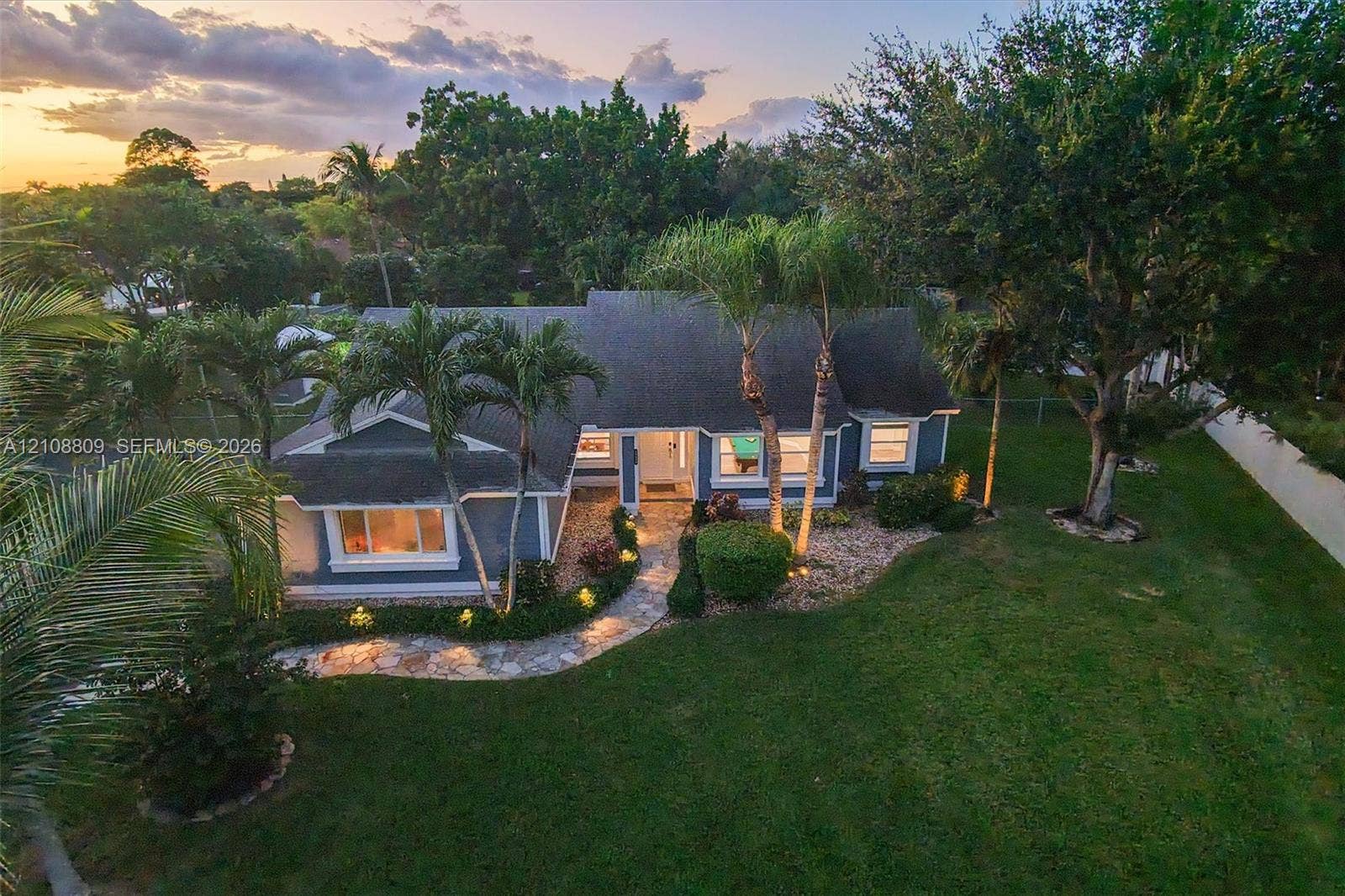 Single-story residence featuring a shingled roof, stone-paved walkway, and expansive lawn with mature trees and palms