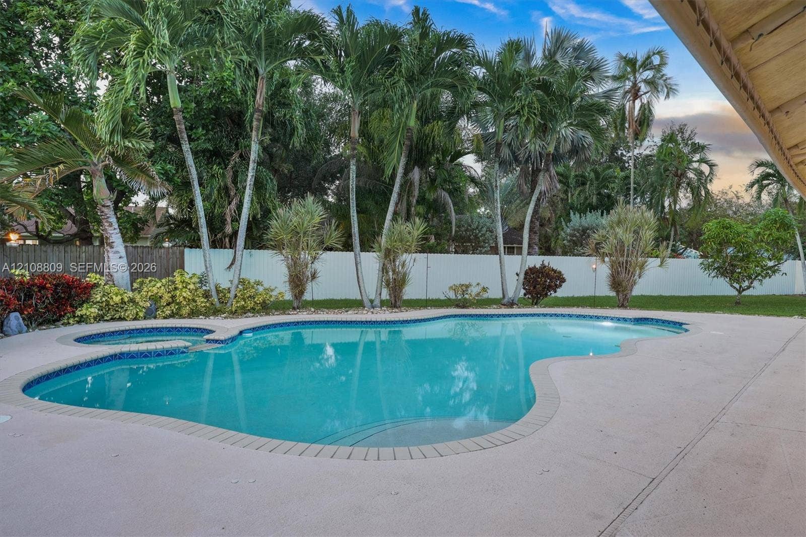 Freeform swimming pool with integrated spa, expansive concrete patio, and perimeter privacy fencing surrounded by mature palm trees