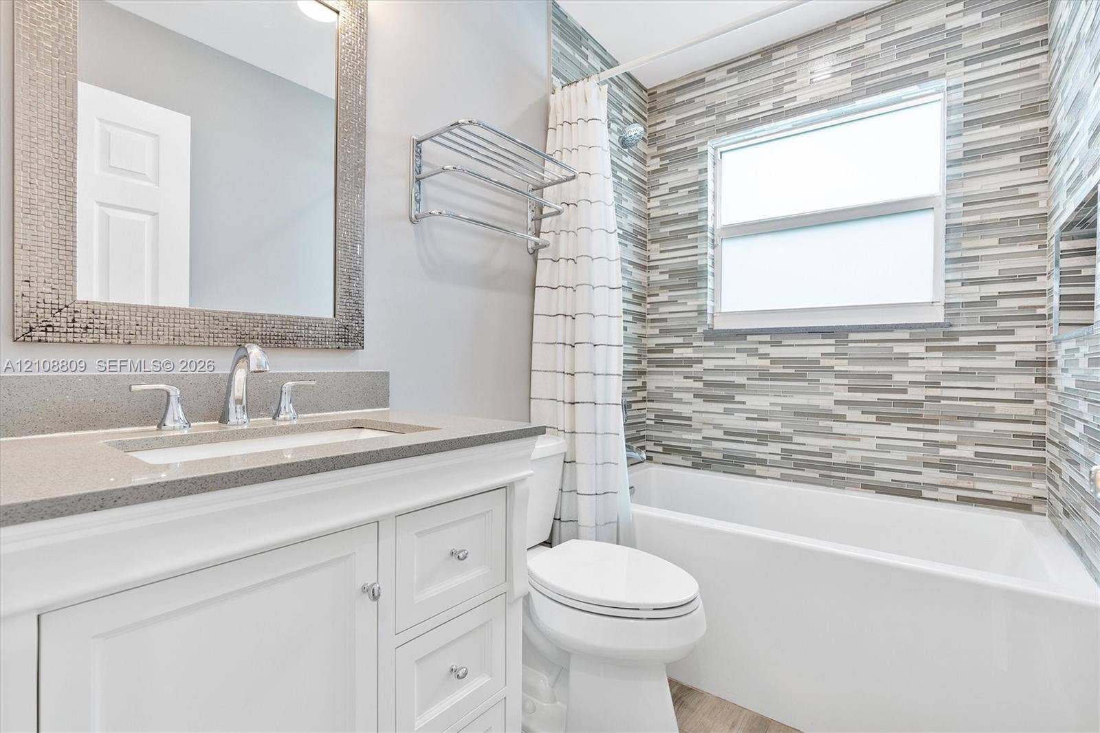Bathroom featuring a white vanity cabinet with a stone-composite countertop, rectangular undermount sink, mosaic-tiled tub surround, and a wall-mounted metal shelving unit