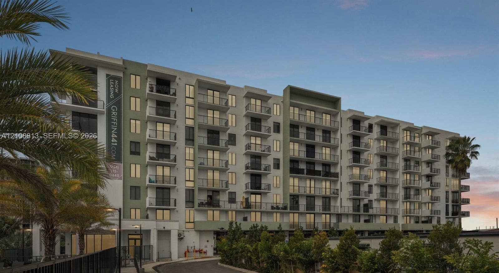 Multi-story residential building featuring a light-toned facade, recessed private balconies with metal railings, and integrated ground-level landscaping