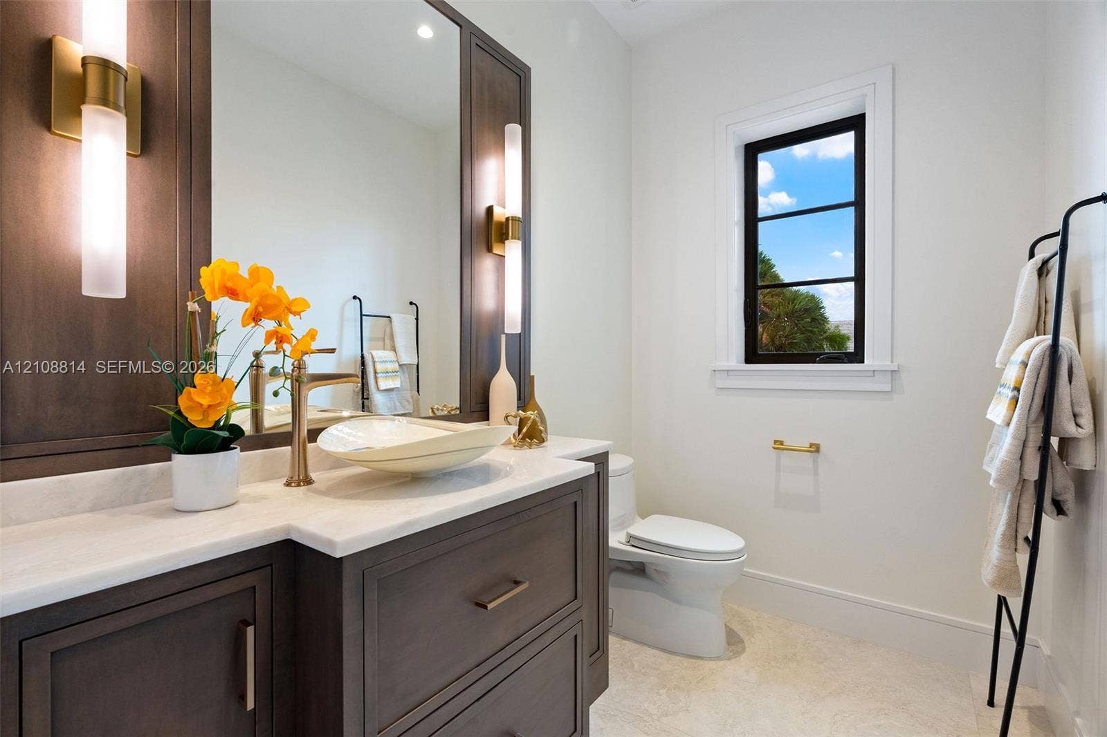 Dark wood vanity featuring a light-toned stone countertop, vessel sink, brass-finish hardware, integrated vertical sconces, and a multi-pane window