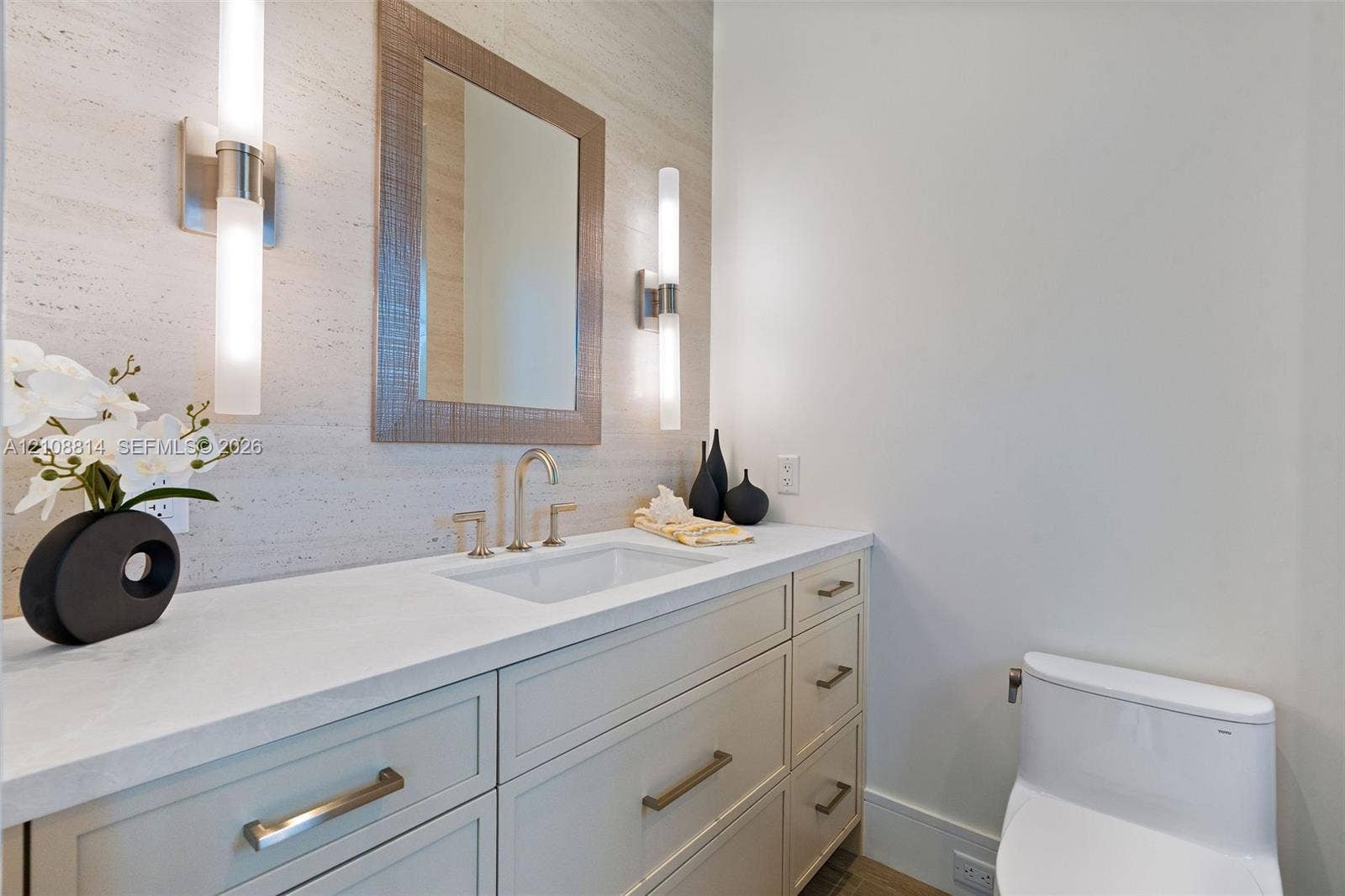 Custom vanity with quartz countertop, undermount sink, stone-finish wall cladding, dual vertical wall-mounted light fixtures, and framed mirror