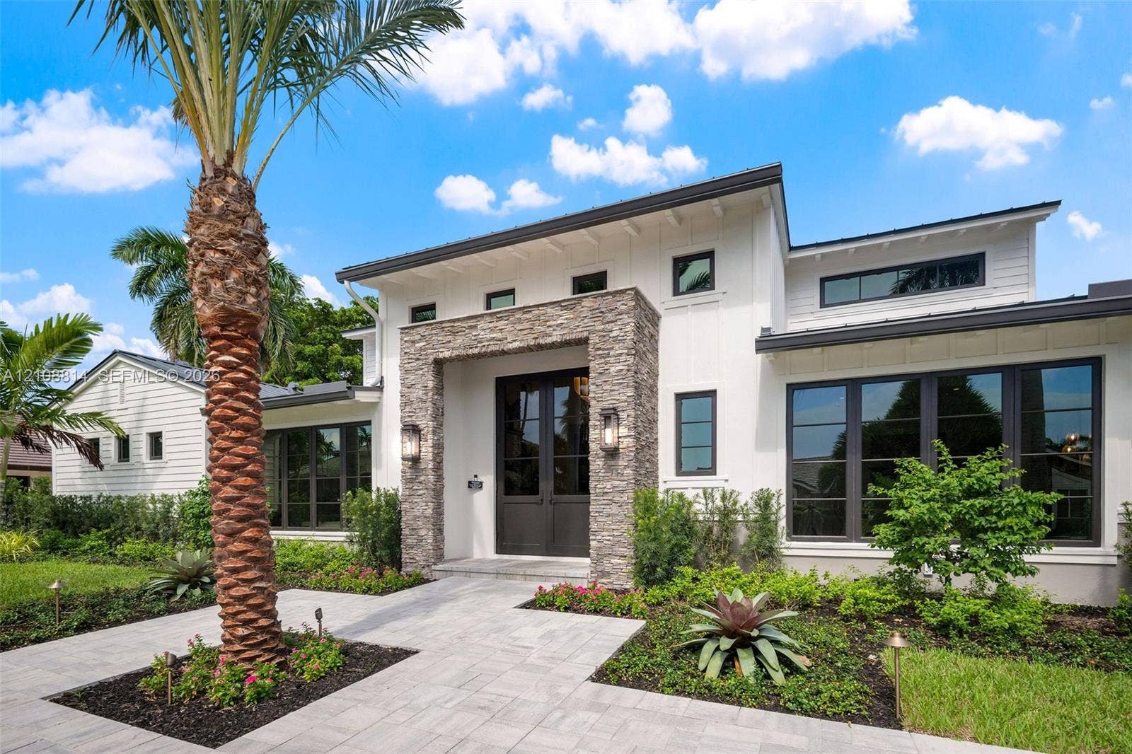 Modern exterior featuring white vertical siding, a stone veneer entryway surround, metal roof accents, expansive dark-framed windows, and a paved walkway