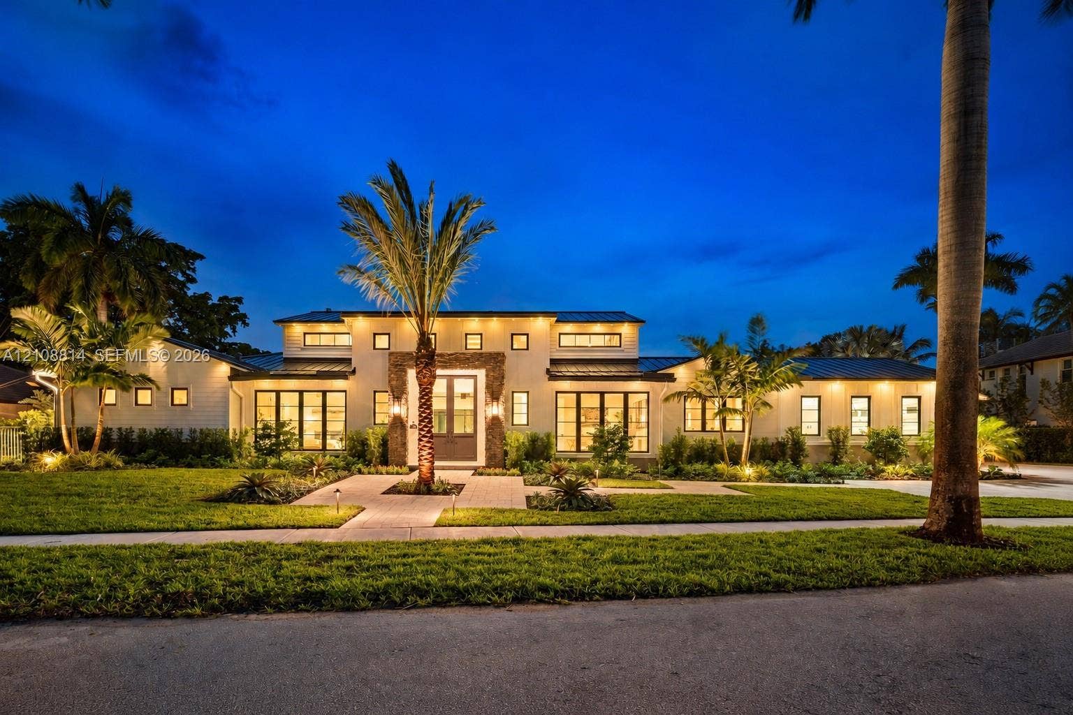Single-story residence featuring light-toned stucco exterior, stacked stone facade accents, dark metal roof, multiple transom windows, and paved stone walkway