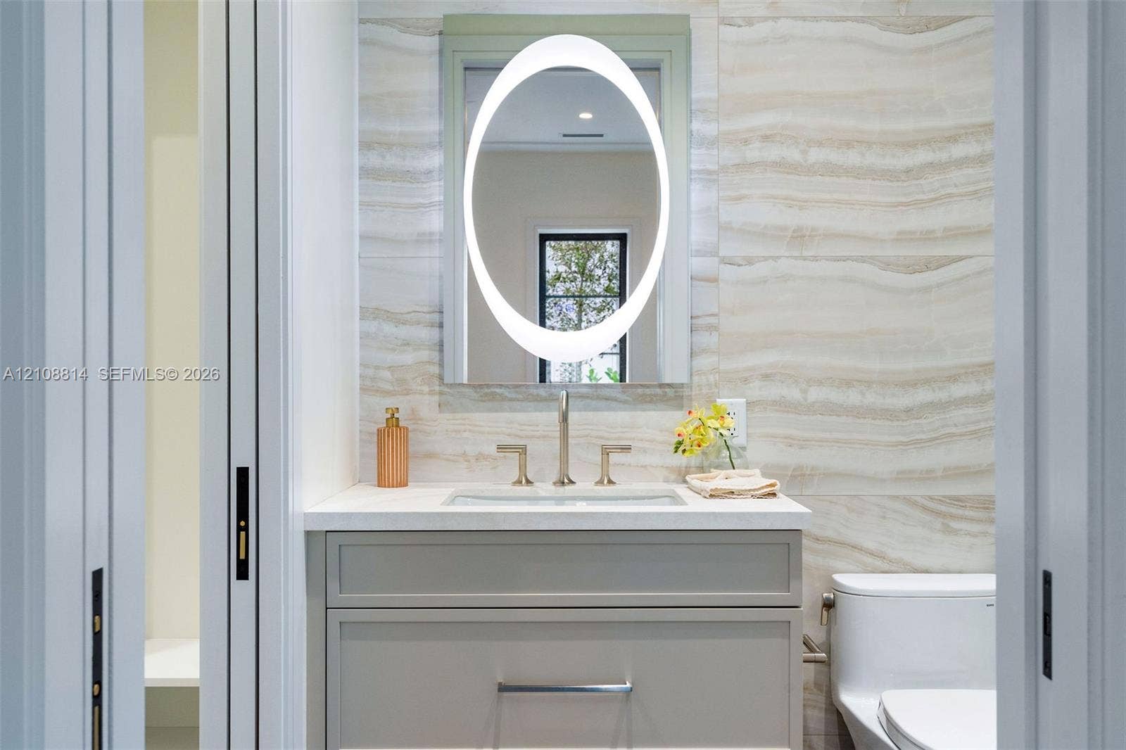 Custom vanity with stone-surface countertop, integrated rectangular sink, oval backlit mirror, and floor-to-ceiling stone-tiled wall